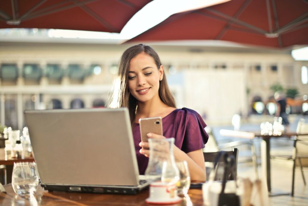 A foto mostra uma mulher, sentada em um café na frente do notebook,  olhando para o celular e sorrindo. Ela ilustra o texto Mundo Pós-Pandemia: sua empresa está pronta para a Era Digital e para o Verdadeiro Século 21?