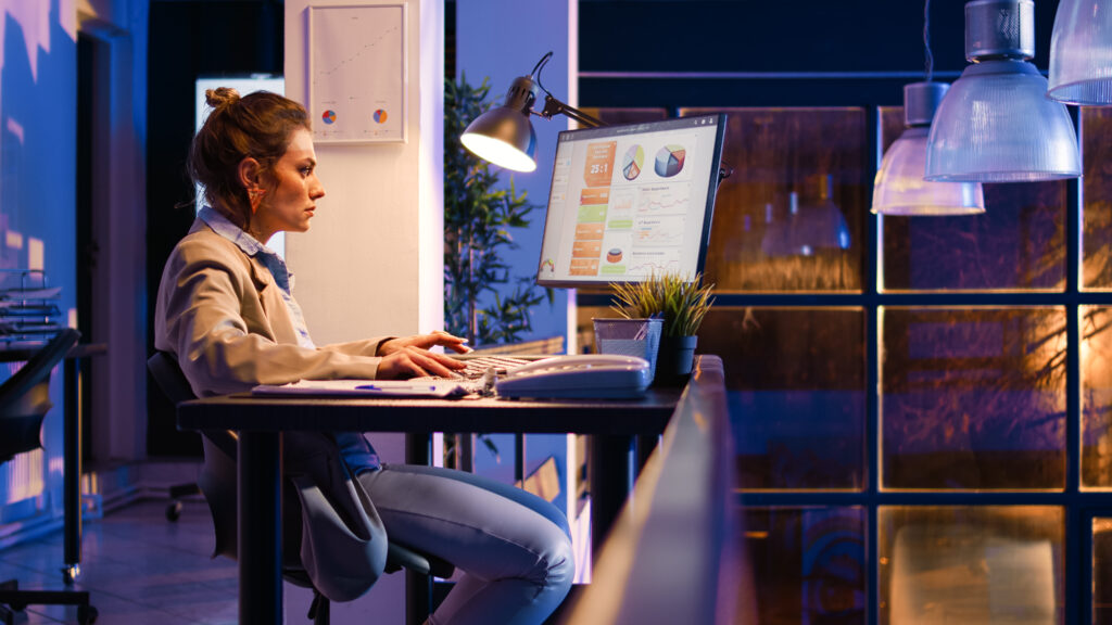 Mulher trabalhando em uma mesa ao computador, olhando gráficos na tela à noite, com uma luz ao lado