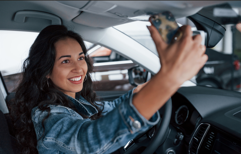 Mulher morena sorrindo dentro de um carro enquanto tira uma foto de si mesma com o celular