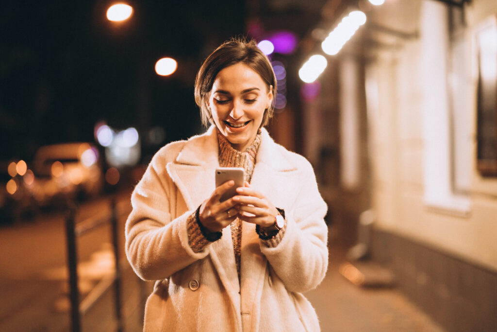 Mulher branca sorrindo olhando para a tela do celular na rua à noite