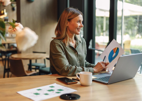 Mulher branca, usando roupa cinza, sorri vendo resultados de marketing em uma mesa de escritório com laptop