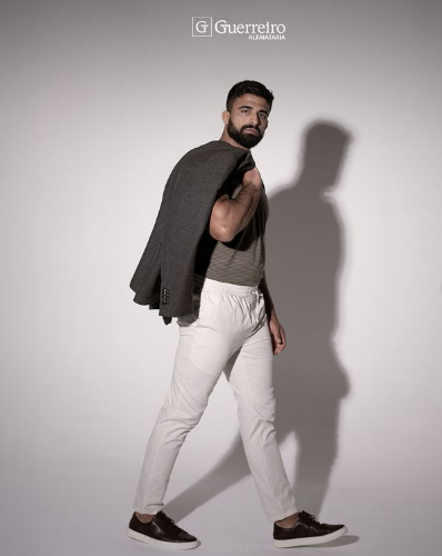 Foto de modelo branco, com barba, posando com uma camisa cinza e calça branca. A sombra dele está projetada na parede